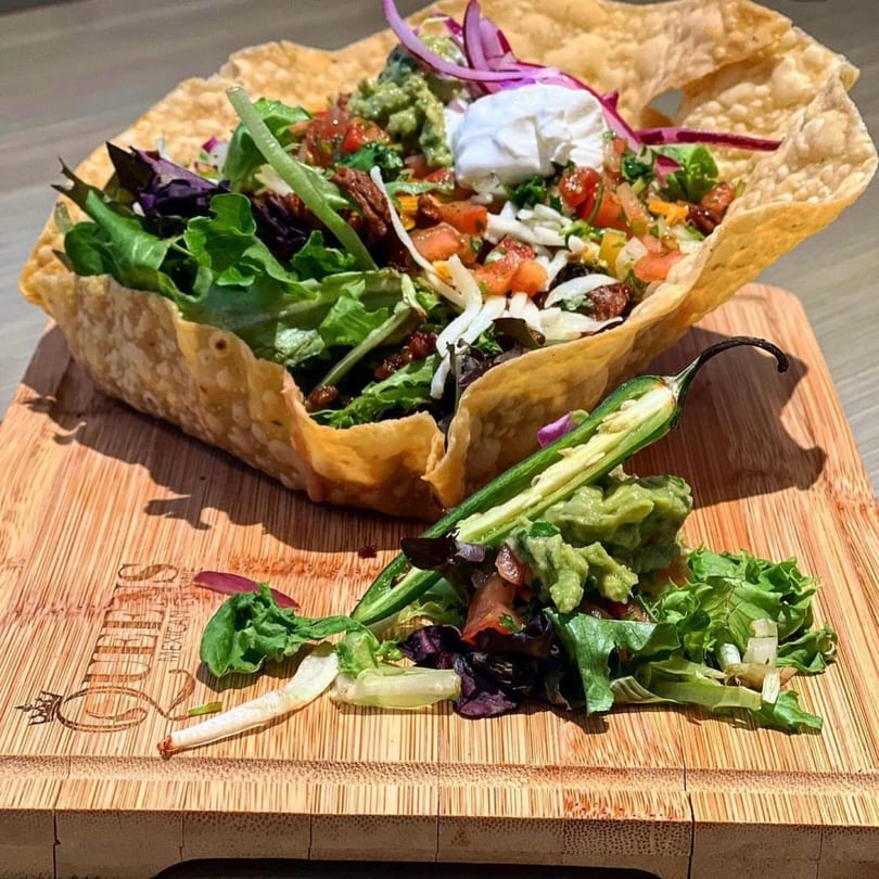 Taco salad in a crispy tortilla bowl with guacamole, sour cream, and a jalapeño on a wooden board.