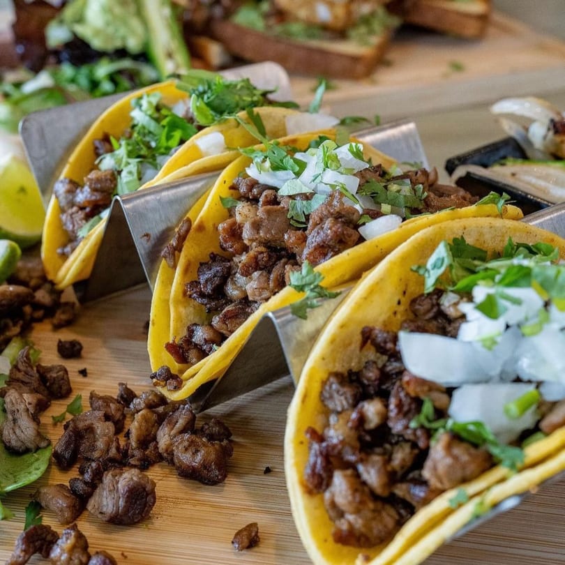 Three beef tacos with cilantro and onions in metal holders on a wooden board.