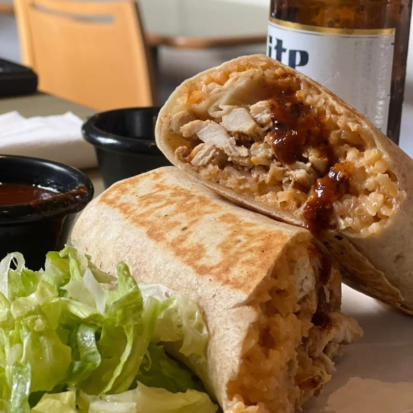 A chicken burrito, cut in half, with sauce, rice, and a side of lettuce, next to a beer.