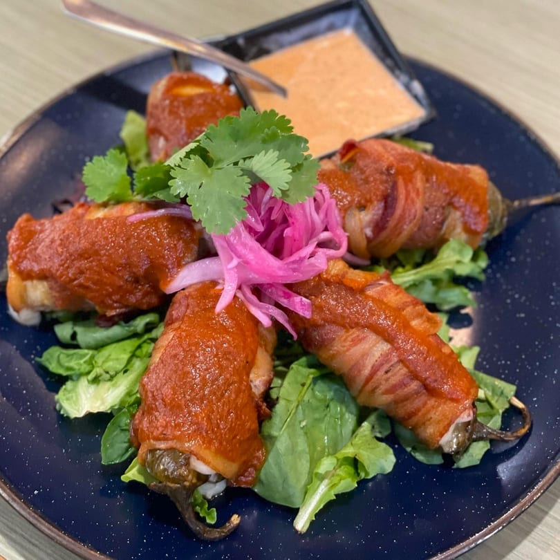 Bacon-wrapped jalapeño poppers with red sauce, pickled onions, and cilantro on a blue plate.