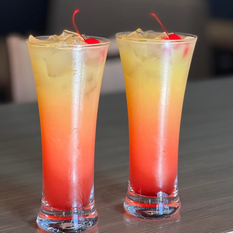 Two vibrant red and yellow cocktails with ice and cherries on a dark wooden table at Queens Bar & Grill.