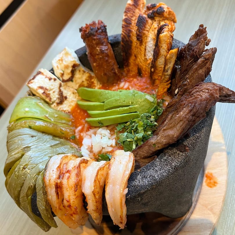 A molcajete filled with grilled meats, shrimp, avocado, cheese, and peppers in a red sauce.