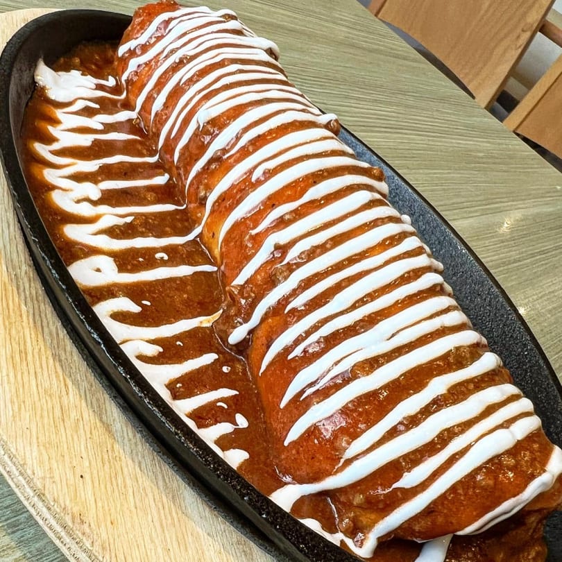 A large, saucy burrito drizzled with white cream, served in a black skillet on a wooden table.